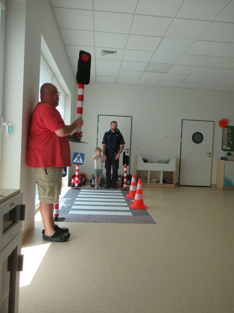 na zdjęciu widać policjanta, dziecko i nauczyciela, oraz mate przejścia dla pieszych